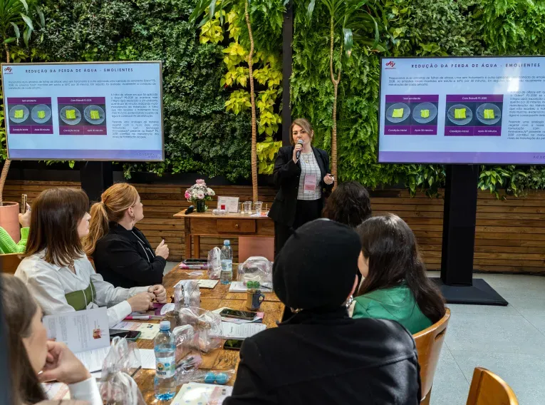 Pessoas sentadam em uma mesa assistindo a uma palestra. Uma mulher está à frente, com um microfone, apresentando slides.