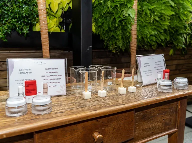 Mesa de madeira com produtos cosméticos e recipientes de vidro para demonstrações. Placas identificam sugestões de formulações como bastão de fragrância, gel foundation e balm.