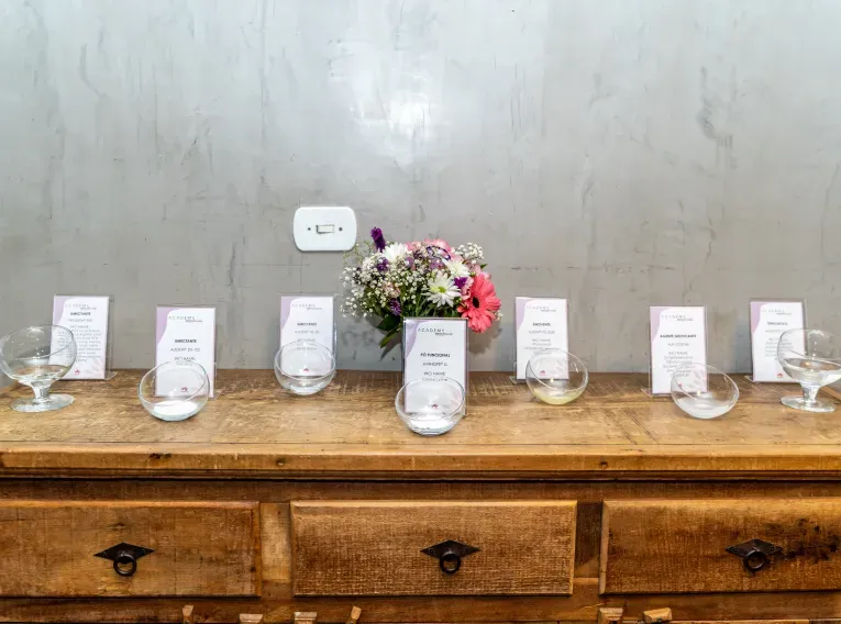 Mesa de madeira com taças de vidro contendo ingredientes cosméticos identificados por placas, como "emoliente" e "agente de textura". Ao centro, um arranjo de flores coloridas.