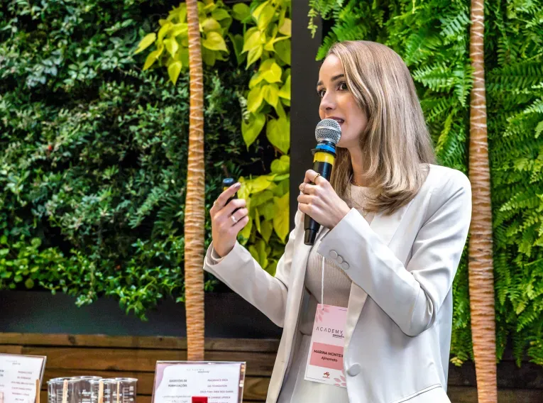 Mulher apresentando em um evento, segurando um microfone e um controle. Ela usa blazer branco e crachá