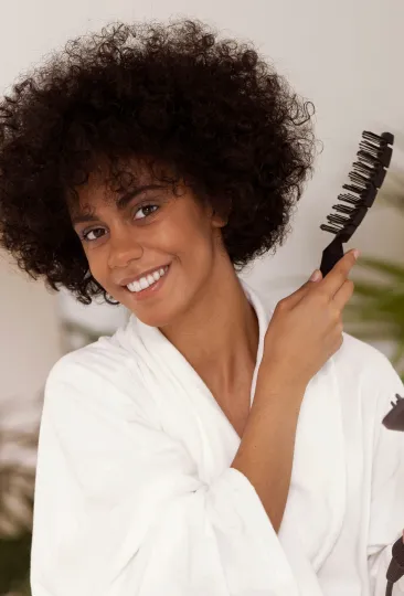 Mulher sorridente em um robe branco enquanto segura uma escova de cabelo, pronta para cuidar dos cabelos volumosos e cacheados.