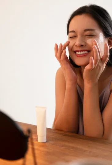Mulher com expressão relaxada, aplicando creme hidratante no rosto, destacando uma rotina de cuidados com a pele em um ambiente iluminado.