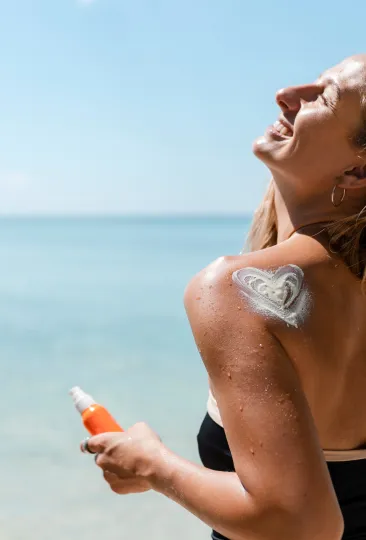 Mulher ao ar livre, em um dia ensolarado na praia, aplicando protetor solar no ombro, onde aparece um desenho em forma de coração feito com o produto.